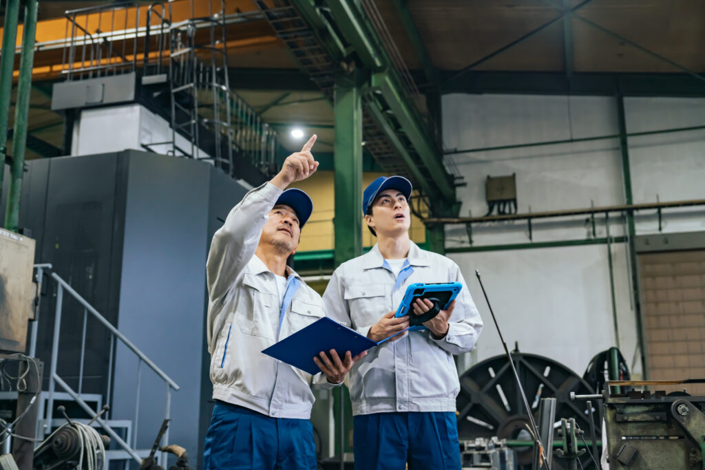 製造業のDX推進担当者とベテラン作業員が協力して生産管理システムの改善を進める工場の様子